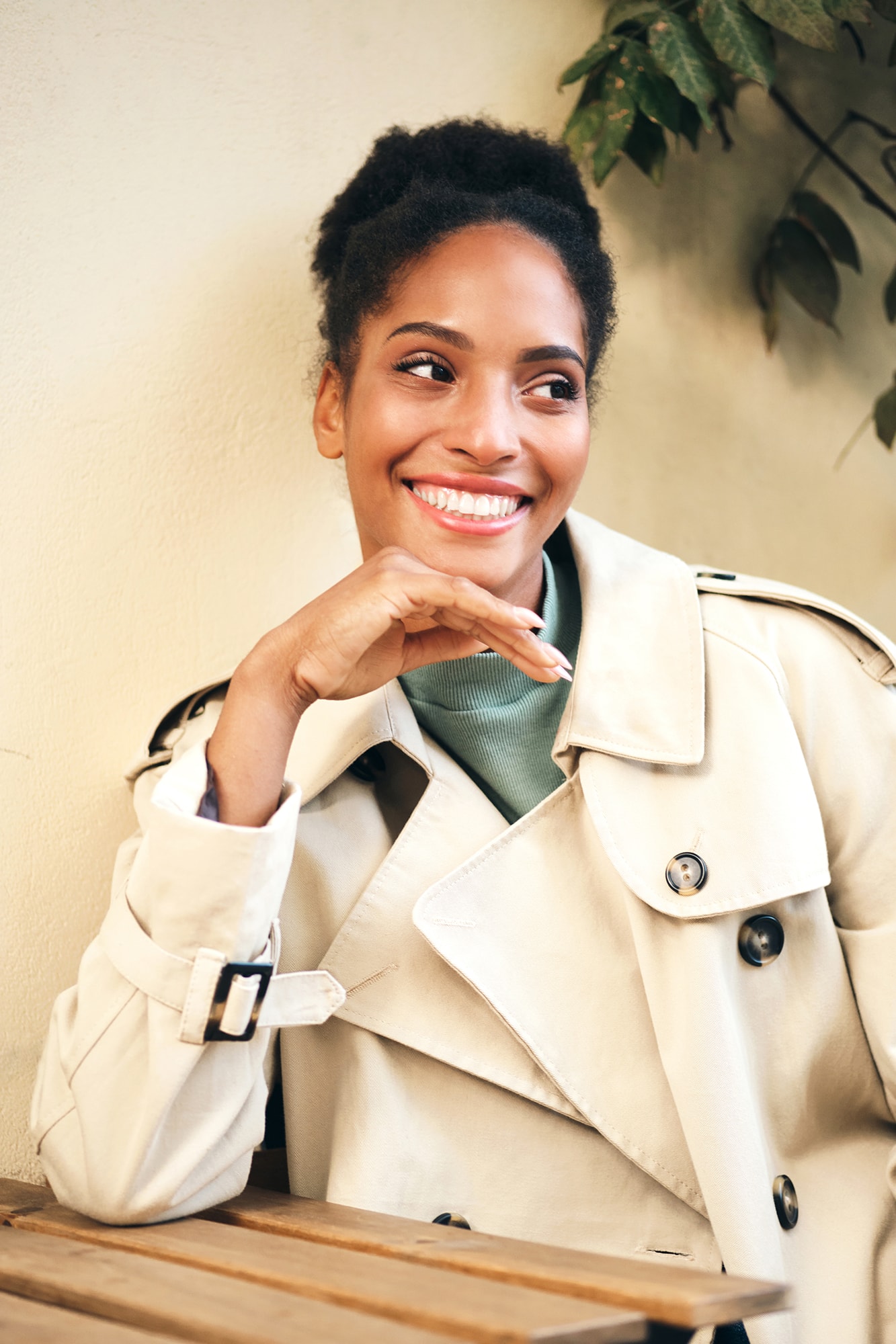 Joyful casual African American girl in stylish trench coat happily looking away in cafe on street