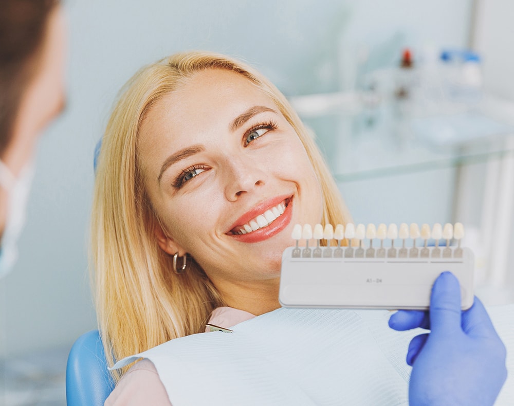 Young man doctor in white gown show veneers enamel color palette for smiling happy patient woman 20s sit at dentist office chair indoor cabinet near stomatologist. Healthcare oral aesthetic treatment.